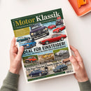 Titelbild der Zeitschrift „Motor Klassik“ mit mehreren farbigen Abbildungen klassischer Autos, darunter Alfa Romeo Giulia, Ford Capri II, MGB GT, Opel Ascona B und Mercedes 180. Überschriften: „IDEAL FÜR EINSTEIGER! 7 Klassiker, mit denen Sie nichts falsch machen können“, „Fotografie-Extra“, „Rolls-Royce Flying Spur & Daimler Double Six“. Zwei Hände halten das Magazin über einer weißen Tischfläche.