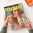 Titelbild der Zeitschrift „myself“ mit einer hellhäutigen Frau in weißem Oberteil und goldfarbenem Schmuck, die ihr Kinn auf die Hand stützt, vor neutralem Hintergrund. Texte: „Mehr Zeit für mich“, „22 Urlaubs-Highlights“, „Haut ohne Hitzestress“, „Das Leben feiern“, „Schluss mit nett“, „Fast wie fliegen“. Oben Titelzeile „Ein Moment für … myself“ in gelber Schrift, gehalten von zwei Händen über hellem Tisch mit orangefarbenem Tablett.