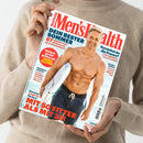 Titelbild der Zeitschrift „Men’s Health“, gehalten von einer Person in beigefarbenem Strickpullover. Auf dem Cover steht ein lächelnder, muskulöser, männlicher Oberkörper in Badehose mit Surfbrett am Strand. Hauptüberschriften: „DEIN BESTER SOMMER“, „EINFACH GENIAL GRILLEN MIT JAMIE OLIVER“, „MIT 50 FITTER ALS MIT 35!“, „VERSTEHST DU DIE FRAUEN?“ sowie Themen zu Fitness, Ernährung und Gesundheit.