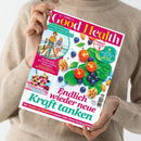 Person in beigefarbenem Strickpullover hält das Magazin „Good Health“ (Ausgabe Juli/August 2023) vor neutralem Hintergrund. Auf dem Cover sind Spinatblätter, Cherrytomaten, Blaubeeren, Walnüsse und Mandeln abgebildet, dazu Titel „Endlich wieder neue Kraft tanken“ sowie Texte zu Themen wie Bewegung, Immunsystem und Ernährung.