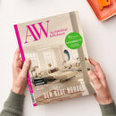 Aufgeschlagenes Magazin „AW Architektur & Wohnen“ mit beigefarbenem Cover, pinker Randgestaltung und Foto eines hellen Wohnzimmers, gehalten von zwei Händen einer nicht erkennbaren Person. Sichtbare Texte: „69 JAHRE RAUM FÜR INSPIRATION“, „36 Seiten Spezial Ferienimmobilien“, „DER NEUE NORDEN“, „Trondheim Norwegens kreative Stadt am Fjord“ und „Architektur & Wohnen“; oben rechts liegt ein orangefarbener Gegenstand auf einem weißen Tisch.