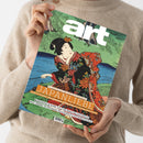 Eine Person im beigefarbenen Strickpullover hält ein schräg ausgerichtetes, glänzend gedrucktes Kunstmagazin. Das Cover zeigt einen japanischen Farbholzschnitt mit einer Frau im blumengemusterten Kimono vor grüner Landschaft und blauem Wasser; sichtbare Texte: „DAS KUNSTMAGAZIN/AUGUST 2026“, „JAPAN LEBEN“, „MIT POSTER-EDITION ZUM HERAUSNEHMEN!“, „Schau in Dresden: Wie japanische Farbdrucke ganz Europa bezauberten“, „MANIFESTA 16 RUHR“ und „PAUL SIGNAC IN POTSDAM“.