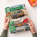 Hochformatige Zeitschrift „Klassik Auto Bild“ auf weißer Fläche, schräg von oben fotografiert und von zwei Händen gehalten. Das Cover zeigt einen beigefarbenen Mercedes mit Dachgepäckträger vor einer Wüstenlandschaft sowie weitere Oldtimer; lesbare Texte sind „DIE JAGD AUF W123“, „Im alten Benz quer durch Marokko“, „Klassiker bis 10.000 Euro“ und „Britische Autoliebe“. Oben rechts liegt ein orangefarbenes Heft oder Tablett.