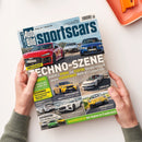 Schräg gehaltenes, glänzendes Automagazin „sportscars Bild Auto“ mit zwei Händen vor hellem Hintergrund; auf dem Cover sind mehrere rote, weiße, blaue, gelbe und grüne Sportwagen abgebildet. Sichtbare Überschriften lauten „TECHNO-SZENE“, „VERGLEICHSTEST“, „Audi RS 5 HYBRID und ELEKTRO Hyundai Ioniq 6 N gegen VERBRENNER BMW M3 Competition“ sowie „HAUSBESUCH bei Alpine in Frankreich“; oben rechts liegt ein orangefarbenes Objekt.