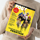 Frau in beigefarbenem Strickpullover hält das Magazin „Tour – Europas Rennrad-Magazin Nr. 1“ mit gelbem Titelblatt in beiden Händen, darauf drei männliche Radsportler in Rennkleidung auf Fahrrädern, mittlerer Fahrer mit erhobenem Arm. Sichtbare Texte: „64 Seiten! Extra-Heft zur Tour de France“, „Tour de France Feiertage“, „Günstige Radhosen für Männer & Frauen“, „32-Zoll-Laufräder“, „Team Telekom“, „Die besten Touren am Arlberg“.