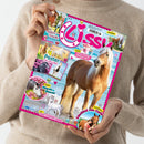 Person in beigem Strickpullover hält eine Ausgabe des Magazins „Lissy“. Titelbild mit großem Foto eines hellbraunen Pferdes mit heller Mähne am Strand, rosa-blauem Schriftzug „Lissy“ und bunten grafischen Elementen. Sichtbare Textaufdrucke u. a.: „4 Poster zum Verlieben“, „Rezepte für deine Einhorn-Party“, „Pferde & Bienen“, „Abschied von Carlo“, „Steckbrief Holsteiner“ und Verlagslogo „Blue Ocean“.