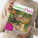 Person in beigem Strickpullover hält das Magazin „GartenFlora“ (Ausgabe August 2026) vor neutralem Hintergrund, Titelzeile „gärtnern, erleben und genießen“. Auf dem Cover: eine Gartenszene mit Holztisch, Bank und bunten Blumentöpfen, Überschriften zu Themen wie „Lieblingsplätze im Garten und auf der Terrasse“, „Sonnengereifte Pfirsiche“, „Kübelpflanzen“ und „Tipps für den Gemüseanbau“.