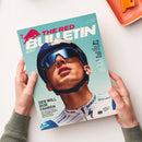 Titelblatt des Magazins „The Red Bulletin“ mit großem Porträt eines männlichen Radsportlers mit weißem Helm und dunkler Sonnenbrille vor hellblauem Hintergrund. Sichtbare Texte: „THE RED BULLETIN“, „BEYOND THE ORDINARY“, „DER WILL NUR FAHREN“, „42 Seiten Sommer-Special: Feel the Real“. Hände in grünem Strickpullover halten das Heft, im Hintergrund eine orange Schale auf weißer Fläche.