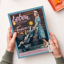 Titelblatt des Magazins „Leben & erziehen“ mit dem Schriftzug „Ferienfeeling“ in großer weißer Schrift und einer grafischen gelben Sonne. Darunter ein Foto einer erwachsenen weiblichen Person im türkisfarbenen Kleid mit weißem Muster und Hut, die einen Kinderwagen schiebt; darin ein kleines Kind. Textinhalte u. a.: „Erfrischung im Glas“, „Schlaf gut, Baby!“, „Urlaub auf Teneriffa“. Hände halten das Magazin über hellem Hintergrund.