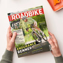 Titelblatt der Zeitschrift „Roadbike“ mit Foto eines männlichen Radfahrers in hellgrünem Trikot und weißem Helm auf einem silbergrauen Rennrad, grüne Natur im Hintergrund. Sichtbare Texte: u. a. „ENDURANCE RENNER“, „KETTENÖLE 13 Schmierstoffe im Test“, „GRAVEL-RACER“, „KAUFBERATUNG“, sowie Markenlogos und Themen zu Rädern, Ausstattungen, Fitness und Testberichten. Hände halten das Magazin über hellem Tisch.