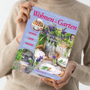 Person in beigefarbenem Wollpullover hält das Magazin „Wohnen & Garten“ Juli 2024 vor neutralem Hintergrund. Titelbild zeigt einen gedeckten Tisch mit weißen Tellern, Gläsern und blauen sowie grünen Blumenarrangements in Metallgefäßen. Sichtbare Texte: „Blau-weiße Sommerträume“, „Dekorieren“, „Feiern“, „Gastlichkeit“, „Garten & Pflanzen“, „Staude des Jahres – Strahlende Schönaster“, „Rezepte mit Blüten & Beeren“.