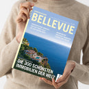 Eine Person im beigefarbenen Strickpullover hält ein schräg zur Kamera ausgerichtetes Magazin mit blau-gelbem Cover. Sichtbare Texte: „BELLEVUE“, „SONDERAUSGABE“, „HANDVERLESENE DOMIZILE – VON DER PROMI-VILLA BIS ZUR KARIBIK-INSEL“, „WOHNTRÄUME ZWISCHEN FRANKFURT, FLORIDA UND FIDSCHI“ sowie „DIE 300 SCHÖNSTEN IMMOBILIEN DER WELT“; das Titelbild zeigt eine Villa am Meer.