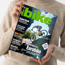 Person im hellbeigen Strickpullover hält ein schräg fotografiertes Magazin mit schwarzem Cover. Darauf stehen „bike“, „Gabel-Duell RockShox ZEB gegen Fox 38 im Hardcore-Test“, „Canyon gegen YT und Radon“, „Kids-Stuff“, „Bikepacking“ und „Die Highlights der Eurobike“; das Titelbild zeigt eine Person auf einem gelben Mountainbike vor felsiger Landschaft.