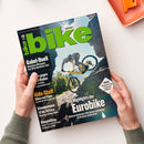 Schräg gehaltenes Fahrradmagazin mit großem grünem Titel „bike“ und dem Text „DEINE NR. 1“, auf dem ein Mountainbike-Fahrer auf gelbem Fahrrad vor felsiger Landschaft abgebildet ist. Sichtbare Überschriften: „Gabel-Duell“, „Kids-Stuff“, „Bikepacking“, „Die Highlights der Eurobike“ und „TARIFA“; zwei Hände einer nicht näher bestimmbaren Person halten das Magazin.