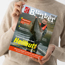 Eine Person ohne sichtbares Gesicht hält ein schräg fotografiertes Angelmagazin „Blinker“ mit blau-grauem Cover und großem weiß-gelbem Titel. Das Cover zeigt einen Angler mit einem großen Fisch am Fluss sowie die Texte „Fliegenfischen“, „Hotspots im Fluss“, „Norwegens spannendster Räuber Heilbutt“, „SPINNFISCHEN LIGHT“ und „NATURKÖDER LIGHT“; im Hintergrund ist ein beigefarbener Strickpullover sichtbar.