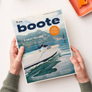 Aufgeschlagenes Magazin „boote“ mit großem dunkelblauem Titel vor einem Berg- und Seemotiv; darunter ein weißes Motorboot mit einer steuernden Person. Lesbare Überschriften sind „Unter Strom!“, „Ruhe im Schiff“, „Reise“, „Boot-Zukunft?“, „Im Test“ und „Stern von Berlin“. Zwei Hände halten das Magazin; oben rechts liegt ein orangefarbenes Heft oder Tablett.