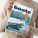 Person ohne sichtbares Gesicht hält mit beiden Händen ein schräg aufgenommenes Magazin „boote“ vor einem beigefarbenen Strickpullover. Das Cover zeigt eine weiße Motoryacht auf blauem Wasser vor felsigen Bergen und ein Segelboot; lesbare Texte sind „Unter Strom!“, „Ruhe im Schiff“, „Boot zaubern?“, „Im Test“, „Reise“ und „Stern von Berlin“.