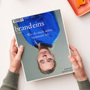 Auf einer weißen Fläche liegt schräg das Magazin „brand eins“ mit dem Titel „Wie du mich siehst, bestimme ich.“ und dem Untertitel „Das geht!“; das Cover zeigt eine weibliche Person mit blauer Gesichtsmaske. Zwei Hände mit grauen Strickärmeln halten das Magazin, rechts oben liegt ein orangefarbenes Kunststofftablett mit orangefarbenem Gegenstand.