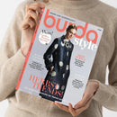 Beige gekleidete Person hält ein schräg fotografiertes Magazin „burda style“ mit hellgrauem Cover und korallenroter Typografie. Darauf abgebildet ist ein weibliches Model in dunkelblauem Mantel mit großen hellen Punkten; lesbar sind „NÄHEN AUS LEIDENSCHAFT“, „Wow! Großes Runway Special“, „HERBST TRENDS!“, „DEEP PURPLE“, „POWER LOOKS“ und „Die schönsten Schnitte der Saison zum Nachnähen“.