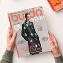 Aufgeschlagene Zeitschrift „burda style“ mit dem Titel „NÄHEN AUS LEIDENSCHAFT“ und dem Foto einer weiblichen Person im dunklen Mantel mit großen hellen Kreisen und Brosche. Sichtbare Texte: „Wow! Großes Runway Special“, „DEEP PURPLE“, „ICONIC FASHION MOMENT“, „POWER LOOKS“ und „HERBST TRENDS! Die schönsten Schnitte der Saison zum Nachnähen“; zwei Hände halten die Zeitschrift, oben rechts liegt ein orangefarbener Gegenstand.