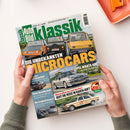Titelblatt der Zeitschrift „Auto Bild Klassik“ mit grün-weißem Logo oben, Hauptüberschrift „Die unbekannten Microcars“ in großen gelb-weißen Buchstaben, darunter Fotos dreier Kleinwagen in Orange, Schwarz und Gelb auf Fahrbahn. Weitere Textfelder kündigen Themen zu Mercedes, BMW, NSU und Opel Manta 400 an. Zwei Hände halten das Heft über weißer Tischfläche mit orangefarbenem Tablett am rechten oberen Rand.