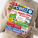 Eine hell gekleidete, hellhäutige Person hält die Computerzeitschrift „PC-WELT+“ in beiden Händen; das Titelblatt zeigt blaue, rote, gelbe und weiße Textflächen mit Schlagzeilen wie „Registry-Tricks mit Sofortwirkung“, „Windows nach Maß“, „Vorsicht vor neuen Phishing-Fallen!“ und „Windows 11 Pannenhilfe“, dazu Symbole für DVDs, Computerbildschirme und Datenträger vor weiß-blauem Hintergrund.
