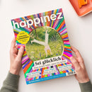 Titelblatt der Zeitschrift „happinez – Das Mindstyle Magazin“, gehalten von zwei Händen über hellem Untergrund. Zentral ein rundes Bild mit einer Person beim Handstand auf einer grünen Blumenwiese, umgeben von einem bunten, strahlenförmigen Muster. Textelemente: „Sei glücklich – Alles strahlt heller, wenn Du lachst“, „Selbst-Erkenntnis“, „Liebes-Magie“, „Aktive Hoffnung“, „Weiblichkeit – Die Superkraft Deines Zyklus“, gelbes Extra-Feld mit „Zauberhafte Lesezeichen“.