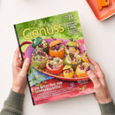 Magazin „Lust auf Genuss“ Nr. 8, gehalten von zwei Händen über hellem Tisch, zeigt auf dem Cover einen rosa Teller mit verschiedenfarbigen, gefüllten Zucchini und Paprika. Textaufschriften: „TOMATEN ZUCCHINI & MEHR“, „Köstlich‑kreativ kombiniert mit Fleisch & Fisch“ sowie „DIE BESTE ZEIT FÜR SOMMERGEMÜSE!“. Grüner Hintergrund mit unscharfem Wiesenmotiv, im Vordergrund leuchtende Gemüse‑ und Servierfarben in Gelb, Grün, Orange und Rot.