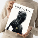 Person in beigefarbenem Strickpullover hält ein Magazin mit weißem Hintergrund und der schwarzen Aufschrift „HOOFORIA – Nachdenken • Bewegen • Verstehen • Begeistern“. Auf dem Cover ist ein schwarzes Pferd mit wallender Mähne in leicht erhobener Pose abgebildet, im unteren Bereich befindet sich ein goldfarbenes „HC“-Logo mit dem Schriftzug „Volume 1“.
