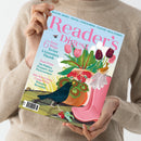 Helle, hellhäutige Person in beigefarbenem Strickpullover hält das Juli-2024-Heft der Zeitschrift „Reader’s Digest“. Das Cover zeigt eine Illustration mit rosafarbenen Gummistiefeln, einem Blumenstrauß aus rosa, weißen und lilafarbenen Tulpen, einer Vase mit roten Blüten, einem schwarzen Vogel und einem kleinen braunen Vogel. Textaufschriften u. a.: „Easy Ways To Get A Green(er) Thumb“, „Sean Penn On Politics…“, „100-Word-Story Competition Winners Revealed“.