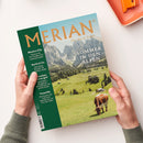 Titelbild des Reisemagazins „MERIAN – The Art of Travel“ mit goldfarbener Schrift; Haupttitel „Sommer in den Alpen“ über einer Berglandschaft mit grünen Wiesen, verstreuten Holzhäusern, einer weidenden braun-weißen Kuh und felsigen Gipfeln im Hintergrund; seitliche grüne Spalte mit Texten zu „Mexico City“, „Korfu & Co“, „München“, „Teneriffa“; gehalten von zwei Händen vor hellem Hintergrund.
