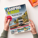 Auf weißem Untergrund liegt schräg eine Zeitschrift, die von zwei Händen einer nicht näher erkennbaren Person gehalten wird; das Cover zeigt einen weißen Camper vor einer mediterranen Landschaft mit Bergen, Zypressen und See. Sichtbare Texte: „CAMPING & REISE“, „MAGAZIN“, „Routen ins Glück“, „Die schönsten Reisen in Europa“, „Spezial 12 Seiten zum Caravan Salon“ und „www.wir-leben-outdoor.de“; oben rechts liegt ein orangefarbenes rechteckiges Objekt.