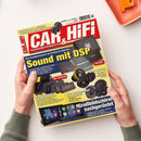 Schräg fotografierte Zeitschrift „CAR&HIFI“ mit rotem, gelbem und blauem Titelblatt, gehalten von zwei Händen; eine Person ist nicht erkennbar. Sichtbare Texte: „Schalldruck gefällig?“, „Sound mit DSP“, „Mini Endstufen“, „Edle Lautsprecher“ und „Großbildschirm nachgerüstet“; abgebildet sind Auto-Lautsprecher, Verstärker und Bildschirme, oben rechts ein orangefarbenes rechteckiges Objekt.