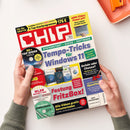 Auf weißer Fläche hält eine Person mit grauem Strickpullover die schräg liegende, farbig gestaltete „CHIP“-Zeitschrift. Das Cover zeigt „Tempo-Tricks für Windows 11“, „SYSTEMSTART + SUCHE + SOFTWARE“, „Festung FritzBox!“, „ARM-Chips: Power pur!“ sowie Abbildungen von Software, Laptop und Datenträgern; oben rechts liegt teilweise ein orangefarbenes Bürogerät auf einer korallenfarbenen Ablage.