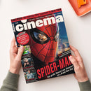 Schräg von oben fotografiertes Film-Magazin „Cinema“, von zwei Händen mit grauen Strickärmeln über einer weißen Fläche gehalten. Das Cover zeigt eine rote Spider-Man-Maske mit schwarzem Netz, den weißen Schriftzug „Cinema“ sowie die Texte „EXKLUSIV: Die coolsten Film-Locations in NEW YORK“, „SPIDER-MAN“ und „Das große Special zum Kinostart von „Brand New Day““; oben rechts liegt ein orangefarbenes Tablett mit einem orangefarbenen Gegenstand.