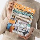 Eine weibliche Person in beigem Strickpullover hält eine schräg fotografierte Zeitschrift mit orangefarbenem Titel „COUCH“ und türkisfarbener Überschrift „FERIENFLAIR ZU HAUSE“. Das Cover zeigt eine Frau in weißer Kleidung mit weißem Hund in einem gestreiften Wohnraum sowie die Texte „URLAUBS-VIBES“, „EUROSUMMER“, „MEERFARBEN“, „ALLES, WAS DU LIEBST“ und „Strandgut upgraden“.