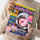 Person mit beigefarbenem Strickpullover hält das Magazin „ZauberTopf – Lieblingsrezepte für Thermomix“ in den Händen; Titelbild zeigt einen Thermomix-Behälter mit rosa Erdbeer-Softeis und Löffel von oben, umgeben von Texten wie „Lust auf Eis mit Thermomix“, „5 Zutaten – frisch & lecker kochen“ und „Nudelsalate“, auf blau-rosa Hintergrund mit gelben und weißen Schriftfeldern.