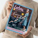 Magazincover „Leben & erziehen“ mit hellblauem Rand; zentrale Abbildung einer Frau in blau gemustertem Kleid und Hut, die einen Kinderwagen schiebt, vor brauner Holz­wand. Sichtbare Texte: Titel „Ferienfeeling“ mit gelber Sonne, Unterzeilen „Erfrischung im Glas – Sommerdrinks für Kids“, „Urlaub auf Teneriffa – Lieblingsziele für Groß und Klein“, „Schlaf gut, Baby! So klappt’s in tropisch­warmen Nächten“ sowie Themenzeile „Ein Fotoshooting macht Mut – Erste Hilfe auf dem Spielplatz“.