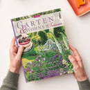 Titelbild der Zeitschrift „Garten & Wohnen Träume“ Ausgabe 3/2026, gehalten von zwei Händen. Das Cover zeigt einen blühenden Sommergarten mit weißer Gartenbank, üppigen violetten und weißen Blumen im Vordergrund sowie grünem Laub im Hintergrund. Textelemente: „Willkommen im Sommergarten“, „Köstliches mit Beeren“, Themen zu Blüten, Gartentouren und Interieur.