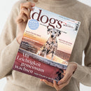 Person in beigem Strickpullover hält Magazin „dogs – Vom Glück, mit Hund zu leben“ in beiden Händen; Titelbild zeigt einen Dalmatiner auf einer rötlich-beigen Mauer vor Sonnenuntergang. Sichtbare Texte: „Mit Leichtigkeit gemeinsam wachsen – Mehr Freude in der Erziehung“, „Hilfe für die Seele“, „Warum tut er das?“, „Gut gemacht!“, „Reif für die Insel – Sardinien mit Hund“ sowie blauer Kreis mit „EXTRA – Die Welt aus der Sicht des Hundes“.