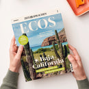 Aufgeschlagenes spanisches Sprachmagazin „ECOS“ von „ZEIT SPRACHEN“ mit dem Titel „Baja California“ und einem Foto von Kakteen, Felsküste, Meer und blauem Himmel. Sichtbare Texte: „SPANISCH“, „GRACIELA ITURBIDE – Ihre Fotos verwandeln den Alltag in Poesie“, „WORTSCHATZ – Vacaciones en el Caribe“ und „Roadtrip in Mexiko von Tijuana bis Los Cabos“; zwei Hände mit grauen Strickbündchen halten das Magazin.