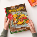Schräg gehaltenes Magazin „Landlust – EIN HAUSGEMACHT!“ mit grünem Rand und Titelfoto von gegrilltem Gemüse, Fleisch und Fisch in einer Auflaufform, von zwei Händen mit grauen Strickärmeln. Lesbare Texte: „Mein Magazin für Haus und Küche“, „Marmeladen“, „Eis selber machen!“, „Schnelles für heiße Tage“, „Gefüllte Fladenbrote“ und „Frisches vom Grill“.