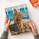 ELLE-Magazin „Deutsche Ausgabe“, von zwei Händen über weißer Fläche gehalten; auf dem Cover sitzt eine blonde Frau mit Muscheloberteil am Strand vor blauem Meer. Sichtbare Texte: „AUGUST 2026“, „LOVE IT! 9 Schritte zu richtig gutem Sex“, „BIG SPLASH – Die besten Hitze-Hacks“, „DAS SONNEN-HOROSKOP – Die Sterne machen Party“, „summer in STYLE“ und „LUFTIGE LOOKS MIT ICED-EFFEKT, COOLE TANKTOPS UND ACCESSOIRES IN SORBET FARBEN“.
