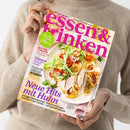 Eine Person in einem beigefarbenen Strickpullover hält ein schräg ausgerichtetes Magazincover mit dem Titel „essen & trinken“ und der Aufschrift „Aus Liebe zum Kochen“. Das Cover zeigt Salat, Tomaten und verschiedene Hähnchengerichte sowie die Texte „Saucen-Schule“, „Zeit für Tomaten“, „Workshop: Die perfekte Pasta alle vongole“, „Italien für Genießer“ und „Neue Hits mit Huhn“.