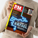 Helle, beige gekleidete Person hält das Magazin „P.M. Das Wissensmagazin“ in beiden Händen; Titelthema „Die Lust am Rätsel“ über einem blauen, leuchtenden Labyrinthmotiv, daneben Schlagzeilen zu „Digitale Forensik“, „Lunar Cruiser“ und „Goldrausch“, oben rechts rotes Feld mit der Aufschrift „Wanted – 5000 Euro Belohnung für den Geistervogel“ und einer Illustration von zwei Vögeln.