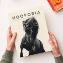 Magazin mit beigem Umschlag und großem schwarz-weiß Foto eines galoppierenden schwarzen Pferdes auf dem Cover, darüber der Titel „HOOFORIA“ mit Unterzeile „NACHDENKEN • BEWEGEN • VERSTEHEN • BEGEISTERN“, unten mittig ein goldenes Logo mit den Buchstaben „HC“ und dem Wort „VOLUME I“; gehalten von zwei Händen mit grünem Strickpullover vor hellem Hintergrund, oben rechts ein orangefarbenes Tablett mit Büroklammern.