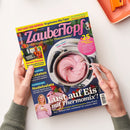 Titelseite des Magazins „ZauberTopf – Lieblingsrezepte für Thermomix“ mit großem Foto eines Thermomix-Behälters, gefüllt mit rosafarbener Eiscreme, daneben ein Löffel in weiblicher Hand. Überschriften u. a.: „Lust auf Eis mit Thermomix!“, „+25 Express-Rezepte“, „Vorrats-Rezepte“, „Nudelsalate“. Farbgestaltung in Pink, Gelb und Blau, Hintergrund hell, Hände halten das Magazin auf weißer Fläche.