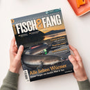 Angelmagazin „FISCH&FANG“ in schräger Draufsicht, von zwei Händen über einer weißen Fläche gehalten; auf dem Cover ein großer Fisch mit grünem Köder vor Flusslandschaft. Sichtbare Texte: „DAS ERLEBNIS-MAGAZIN FÜR ANGLER“, „FISHING WORLD + Filme!“, „Meeresangeln“, „Allein am Wasser“, „Fischküche“, „Trotting am Fluss“, „Rund ums Dekor“, „Heißes Deadbaiten“ und „Alle lieben Würmer“.