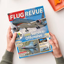 Zeitschrift „FLUGREVUE – Das Luft- und Raumfahrt-Magazin“ auf weißer Fläche, von zwei Händen mit grünen Strickärmeln gehalten. Das Cover zeigt ein Passagierflugzeug und nennt „Königliche Service-Offensive“, „Jordaniens neue Flotte“, „F-35 in Italien“, „Boeing 777-9“ sowie „Helis im Härtetest“.