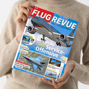 Schräg gehaltenes, blau-rotes „FLUG REVUE“-Magazin aus glänzendem Papier mit einem Passagierflugzeug auf dem Titelbild; darunter Fotos eines F-35-Kampfjets, eines Hubschraubers und eines Flugzeugtriebwerks. Lesbare Texte: „Das Luft- und Raumfahrt-Magazin“, „Königliche Service-Offensive“, „Jordaniens neue Flotte“, „F-35 IN ITALIEN“ und „SPECIAL Helikopter“; zwei Hände und ein beiger Strickpullover, das Gesicht der Person ist nicht sichtbar.